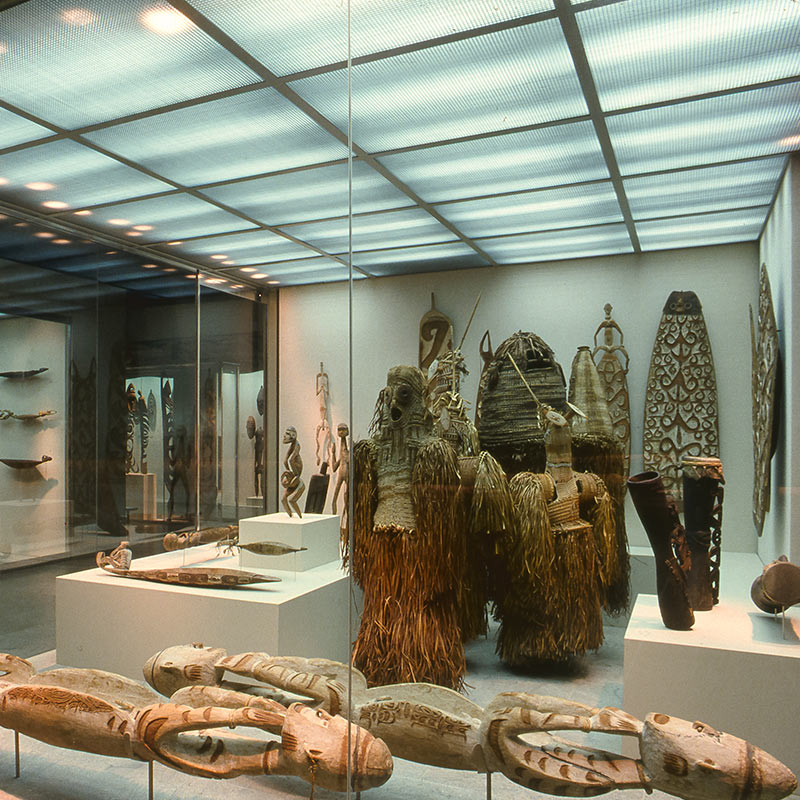 A room-sized glass display with bluish white lights displays various earthy-colored objects. In the middle stand three masks made from palm leaves attached to body-sized suits with stringy, papery brown fiber strings like parchment confetti streaming down to the floor. Behind them are wooden figurines and a large wooden board like a surfboard etched with ornate and loopy engravings.